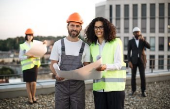 Portrait of caucasian foreman in hardhat and african american inspector in vest looking at camera and smiling on roof. Businessman in suit talking on smartphone while his colleague looking at plan.