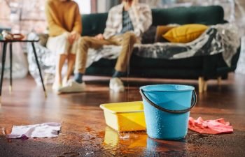 Roof is Leaking, Pipe Rupture at Home: Water Drips into Buckets in Living Room. Angry Couple in Background Calling Insurance Company, Screaming into Phone in Frustration, Trying to Find Plumber